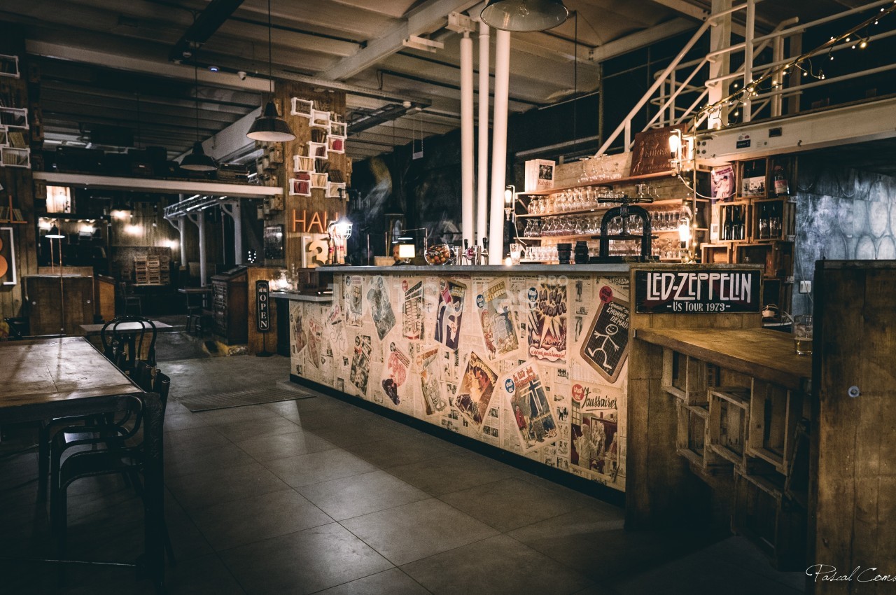 Intérieur d'un bar au style industriel, avec un comptoir décoré d'affiches et une ambiance tamisée.