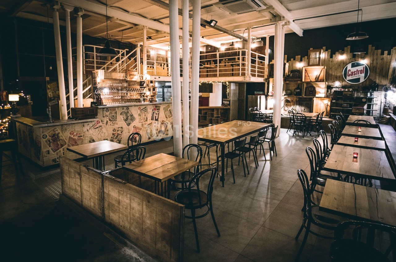Salle de restaurant au style industriel avec tables en bois, chaises noires, bar en arrière-plan et éclairage tamisé.