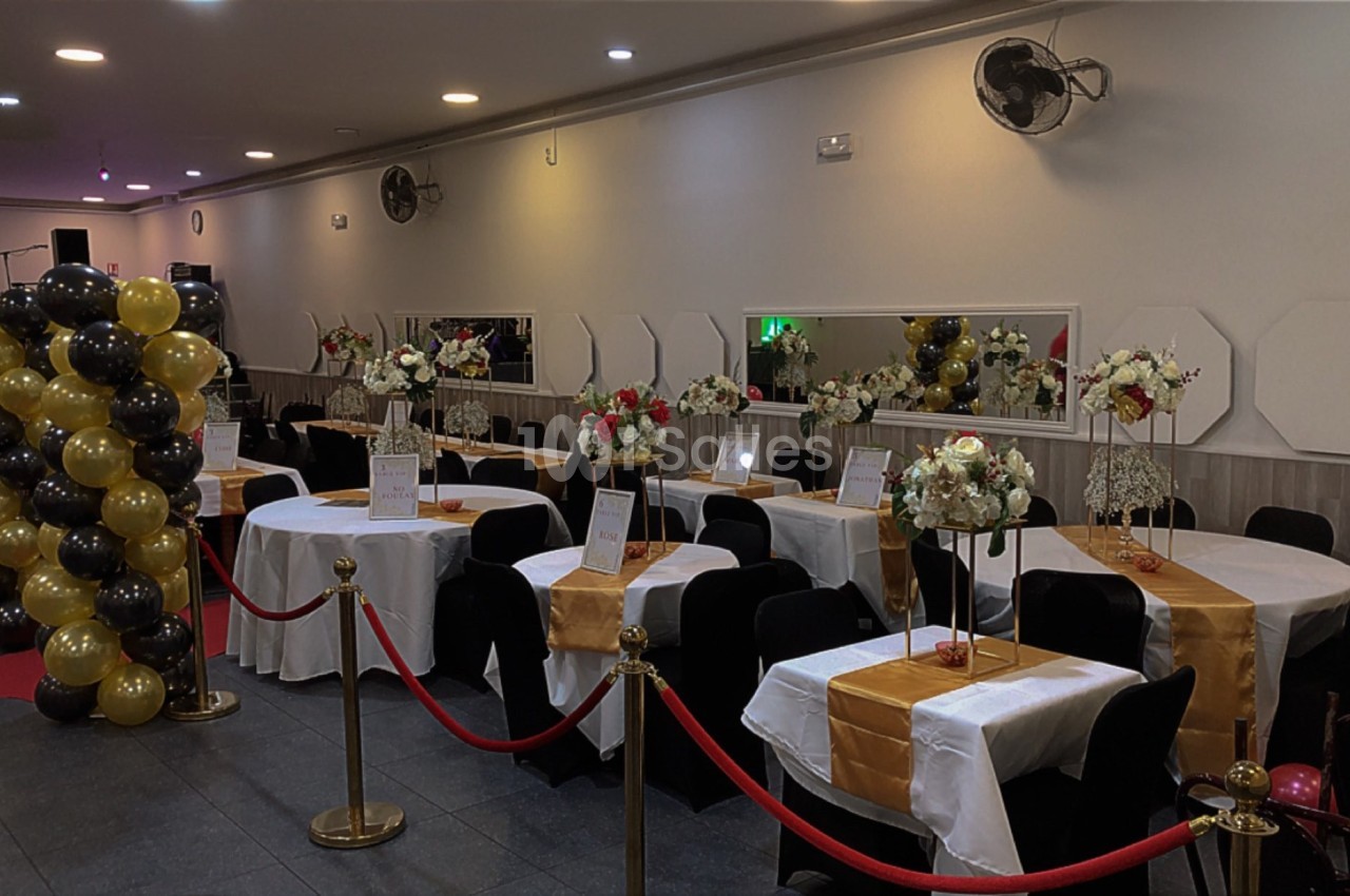 Salle de réception décorée avec des tables dressées, des ballons noirs et dorés, et des fleurs en centre de table.
