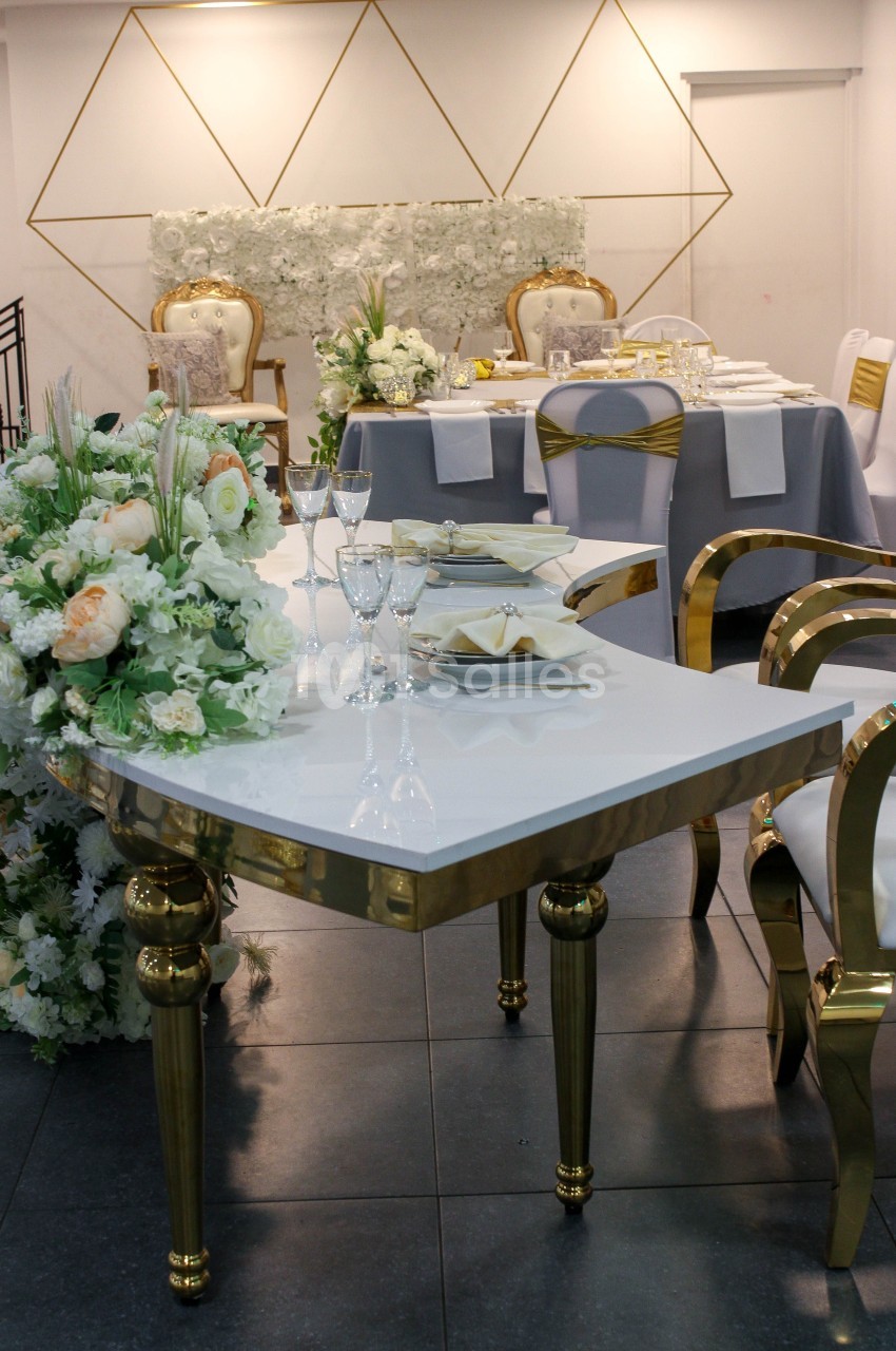 Salle de réception élégante avec tables décorées de fleurs blanches et dorées, vaisselle raffinée et chaises dorées.