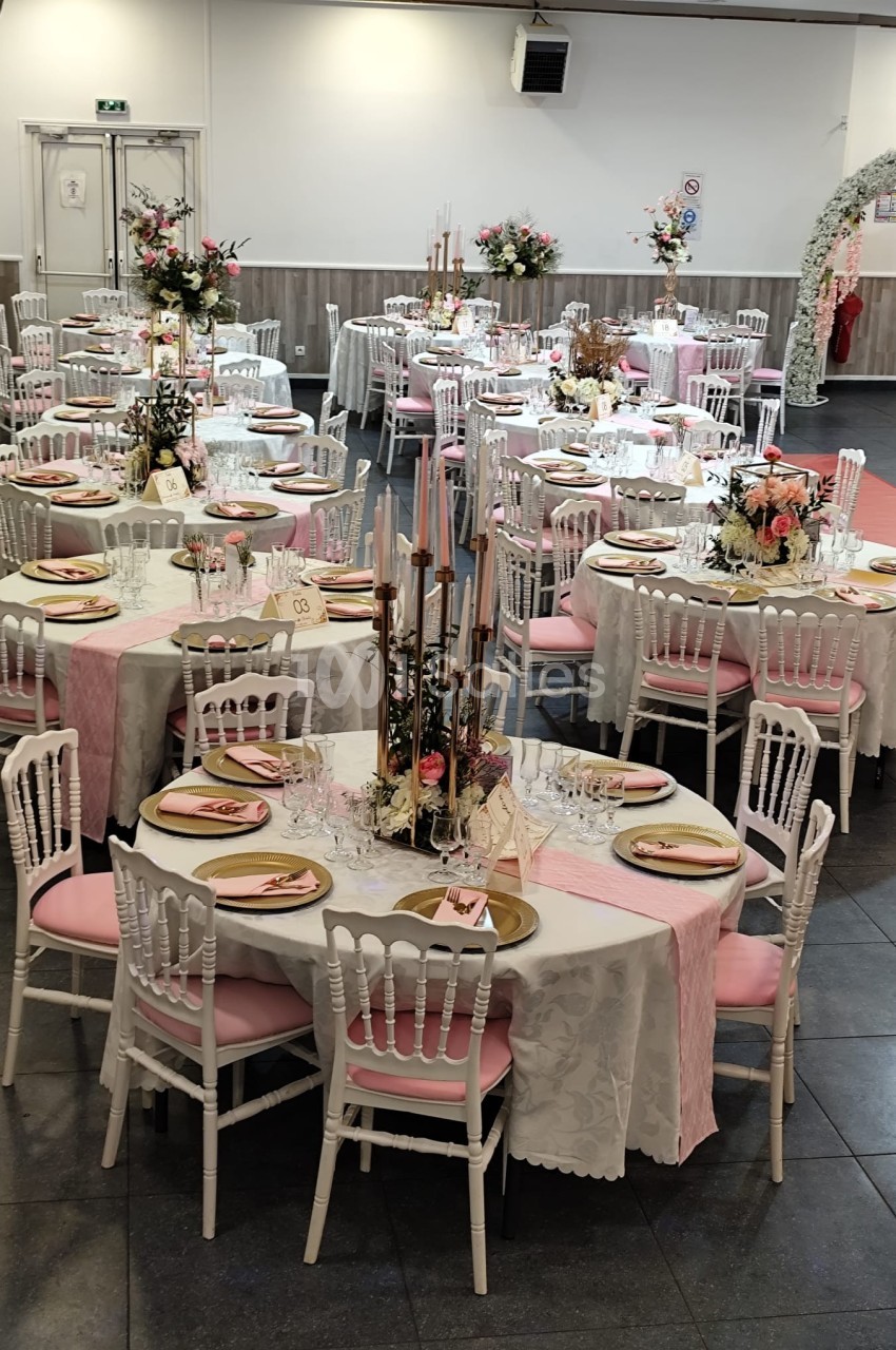 Salle de réception décorée avec des tables rondes ornées de nappes blanches, chemins de table roses et compositions florales.