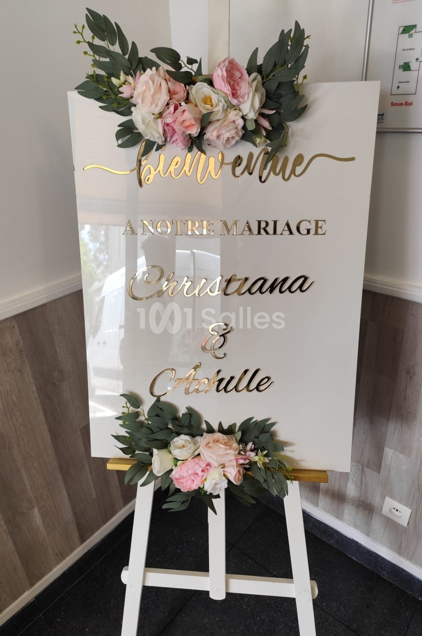 Panneau blanc décoré de fleurs roses et feuillages, portant l'inscription ’Bienvenue à notre mariage’.