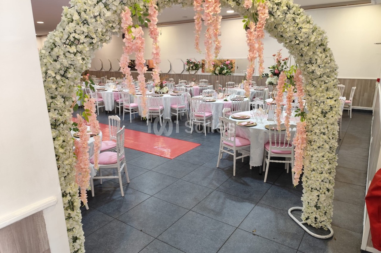 Arche florale blanche et rose menant à une salle de réception décorée avec des tables dressées et des chaises blanches.