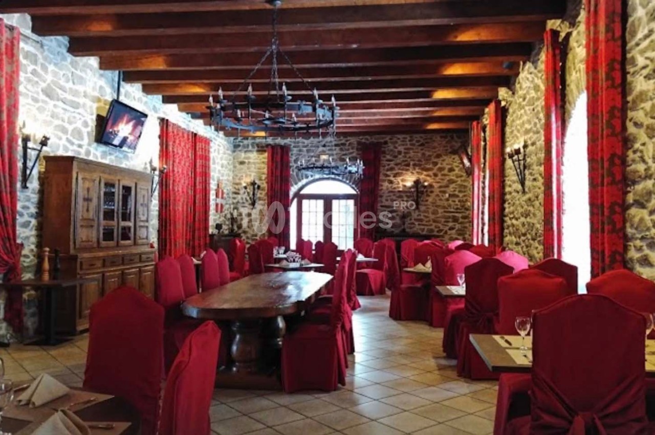 Salle de restaurant avec murs en pierre, chaises rouges, grandes tables en bois et lustre en fer forgé.