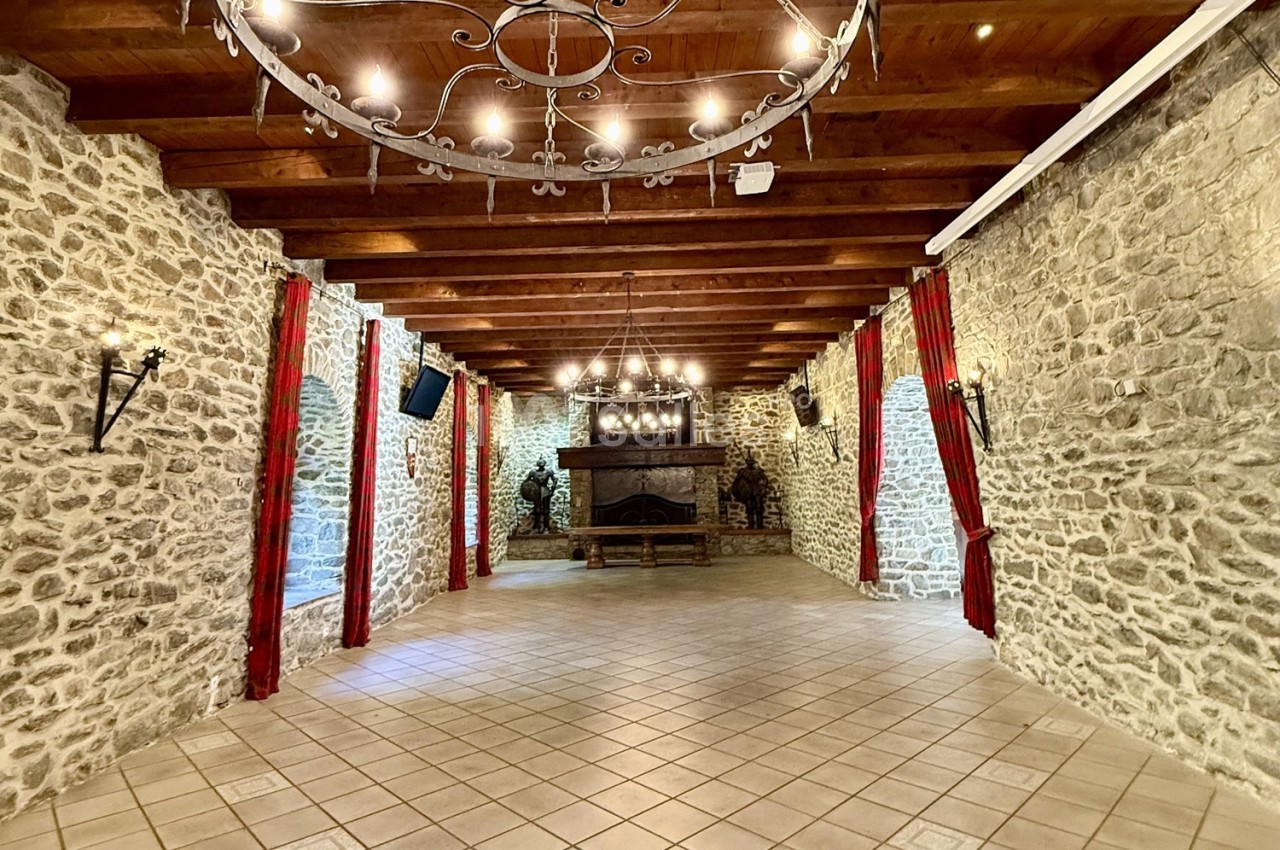 Salle en pierre avec poutres apparentes, cheminée centrale, lustres en fer forgé et rideaux rouges encadrant les murs.