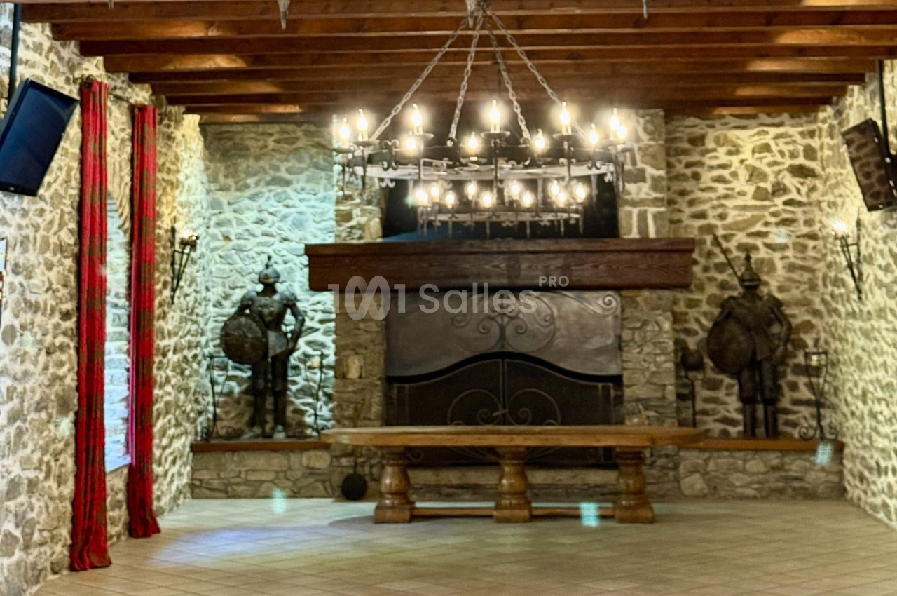 Salle en pierre avec cheminée, banc en bois massif, lustre en fer forgé et statues d'armures médiévales.