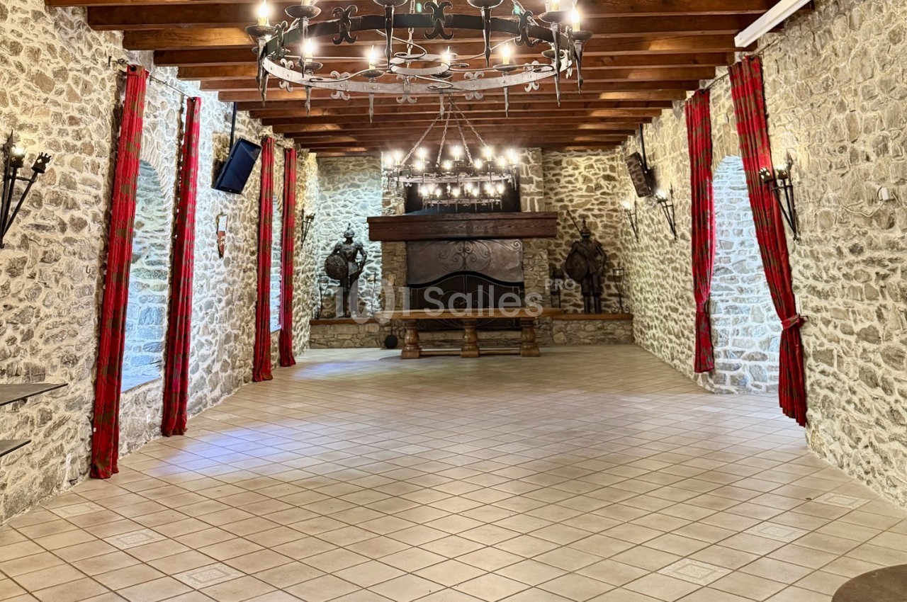 Salle en pierre avec poutres apparentes, lustres en fer forgé, rideaux rouges et grande cheminée en fond.