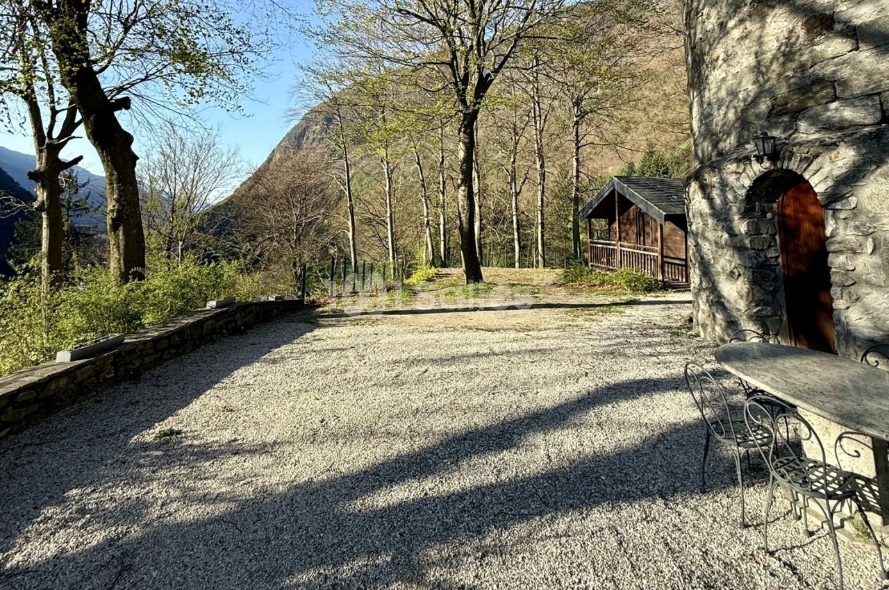 Cour en gravier bordée d'arbres, avec une table en fer forgé, une maison en pierre et un chalet en bois en arrière-plan.
