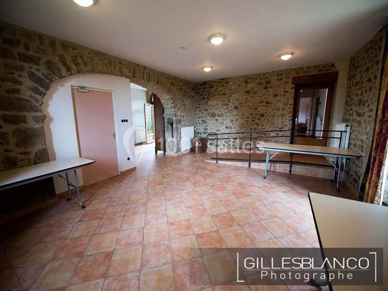 Salle vide avec murs en pierre, sol en carrelage et tables pliantes disposées le long des murs.