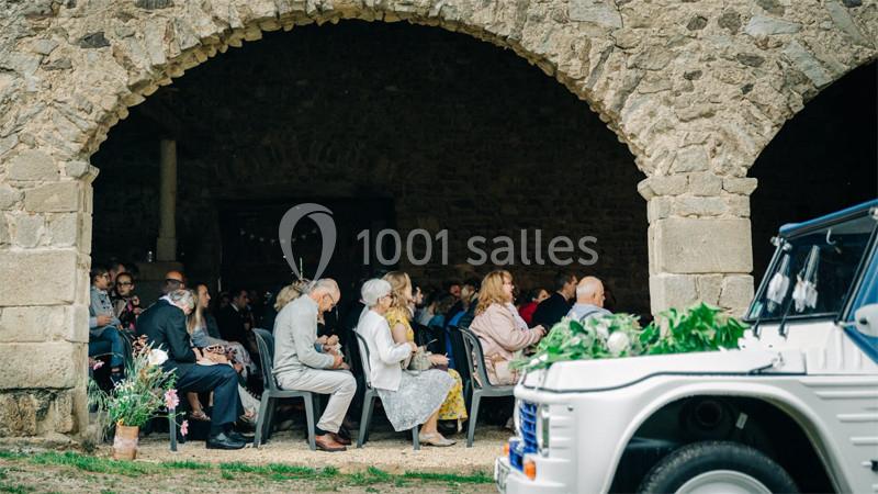 Des invités assis sous une arche en pierre assistent à une cérémonie, avec une voiture décorée au premier plan.