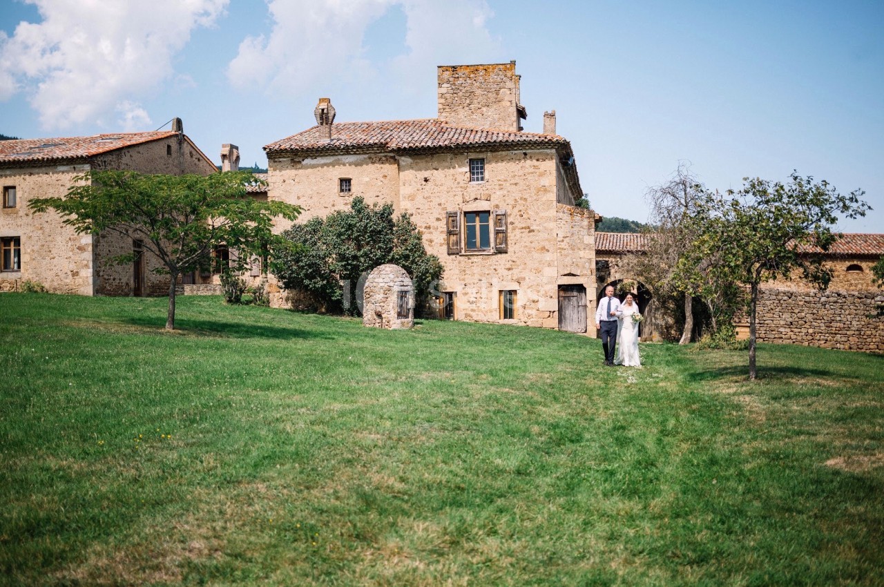 Un couple marche dans un jardin verdoyant devant un bâtiment en pierre ancienne sous un ciel partiellement nuageux.