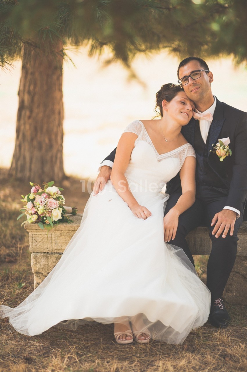 Un couple en tenue de mariage assis sur un banc en pierre, entouré de végétation et d'un bouquet de fleurs posé à côté.