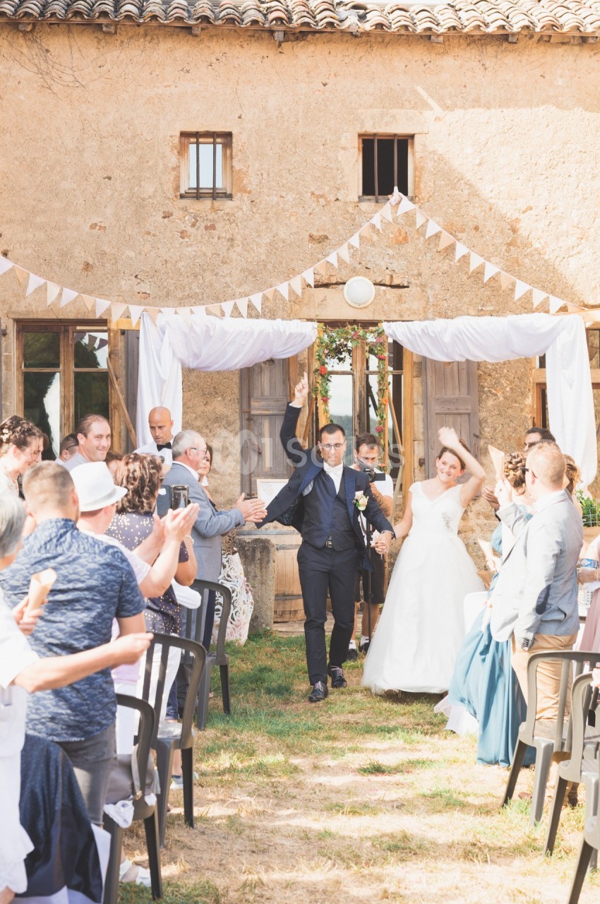 Un couple de mariés souriants sort sous les applaudissements des invités devant une bâtisse en pierre décorée.
