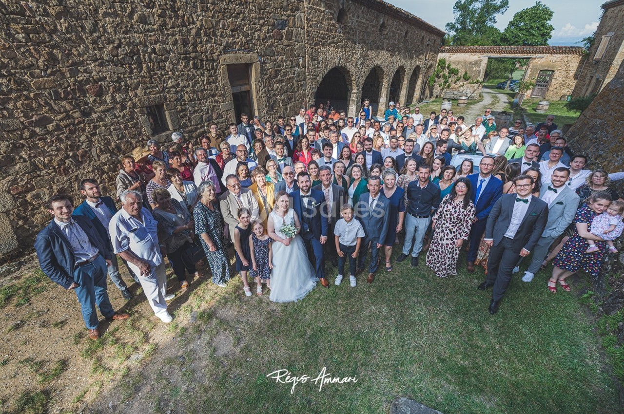 Groupe de personnes rassemblées en extérieur devant des bâtiments en pierre, avec un couple en tenue de mariage au centre.