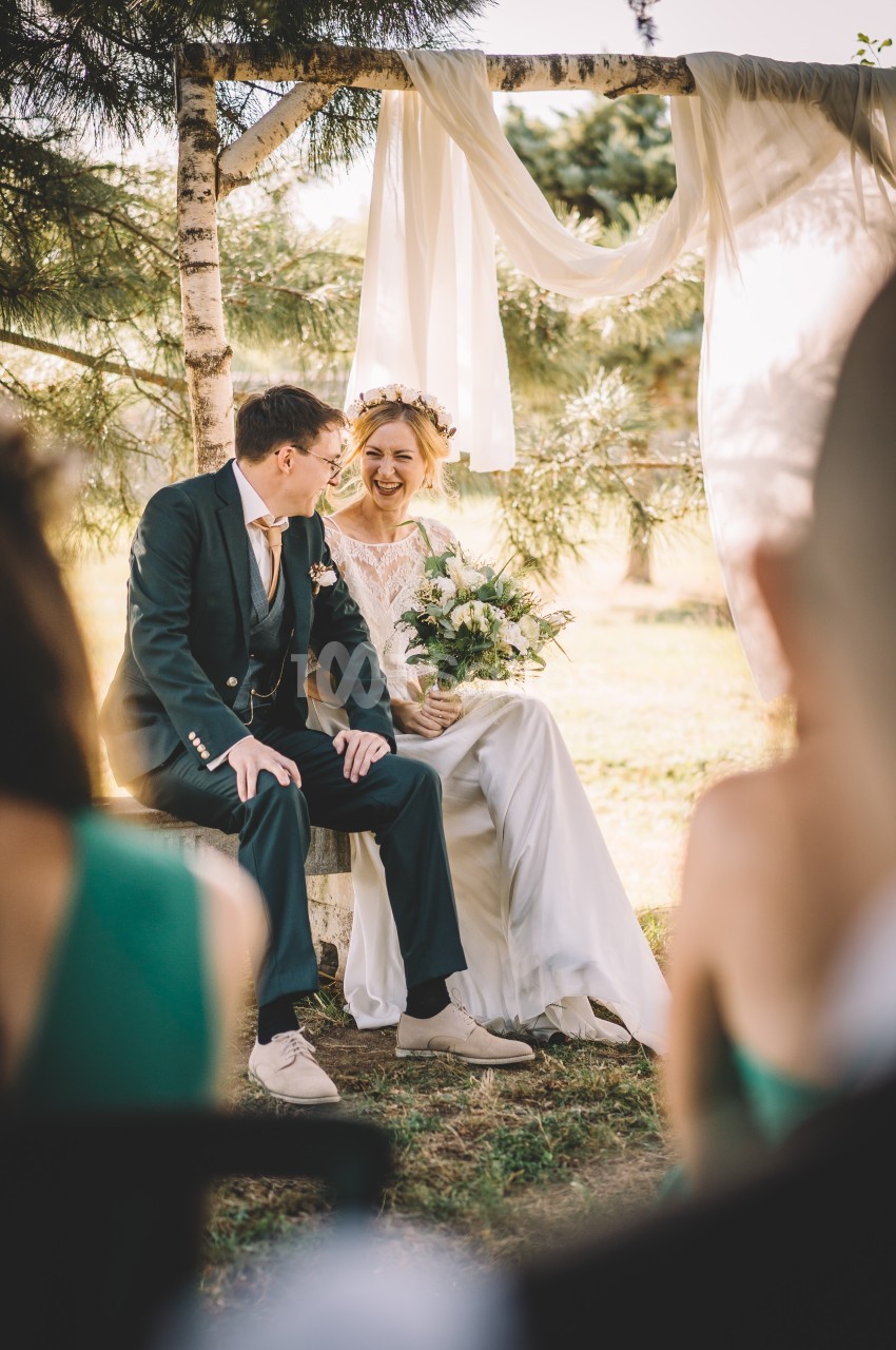 Un couple assis sur un banc en extérieur lors d'une cérémonie, entouré de verdure et de décorations en tissu blanc.