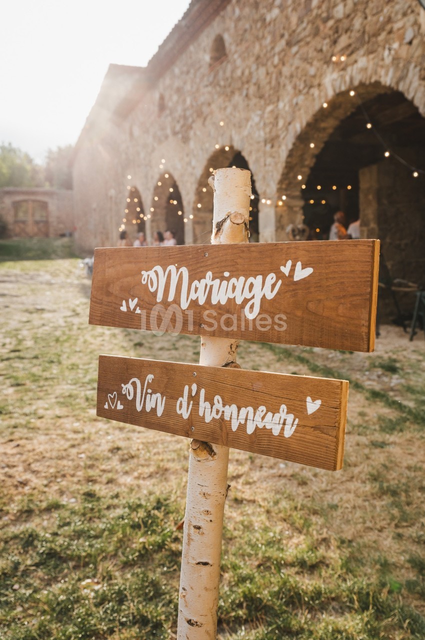 Panneaux en bois indiquant ’Mariage’ et ’Vin d'honneur’ devant un bâtiment en pierre avec guirlandes lumineuses.