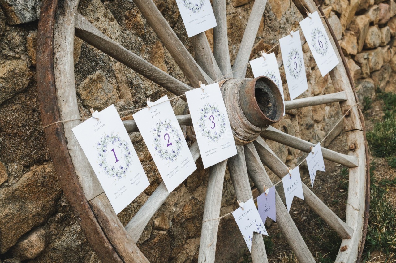 Vieille roue en bois décorée de cartes numérotées et de fanions, posée contre un mur en pierre.