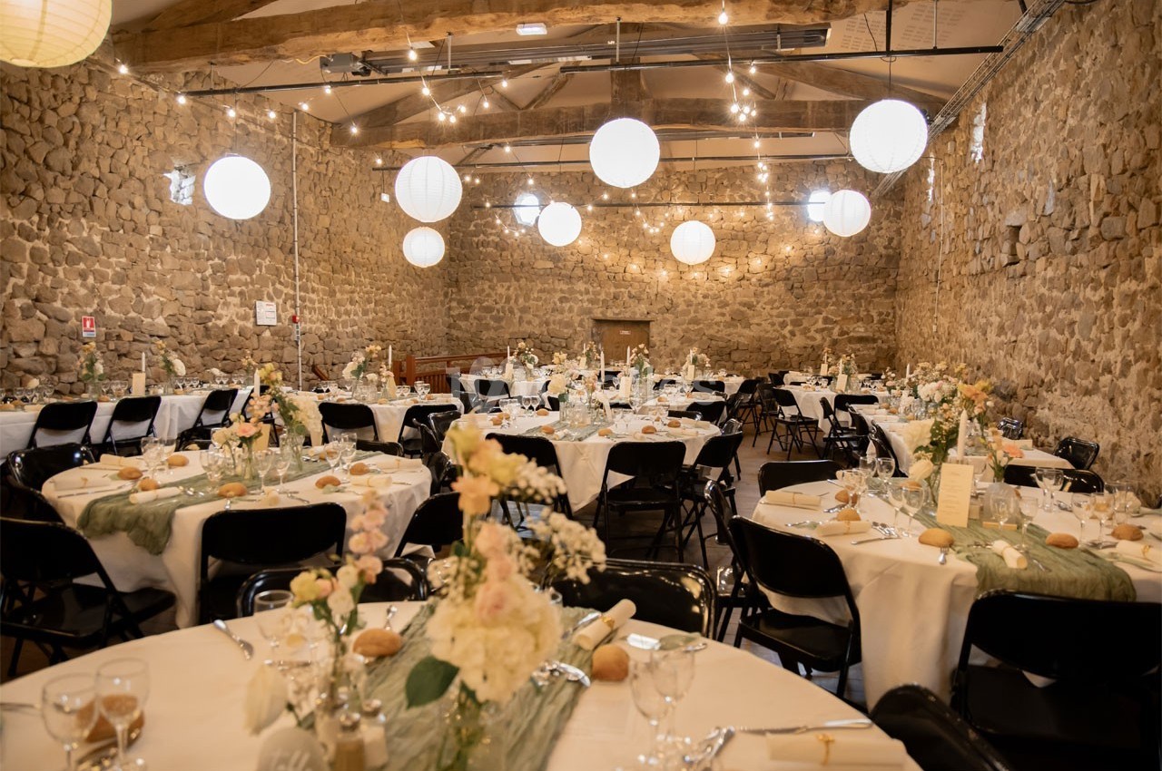 Salle en pierre décorée pour un événement, avec des tables rondes dressées, des fleurs et des guirlandes lumineuses…