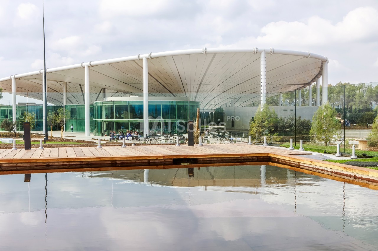 Bâtiment moderne avec une structure circulaire vitrée, entouré d'un bassin et d'espaces verts.