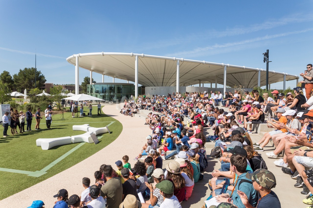 Foule assise en plein air devant une scène, sous un ciel dégagé, près d'un bâtiment moderne avec toit blanc.