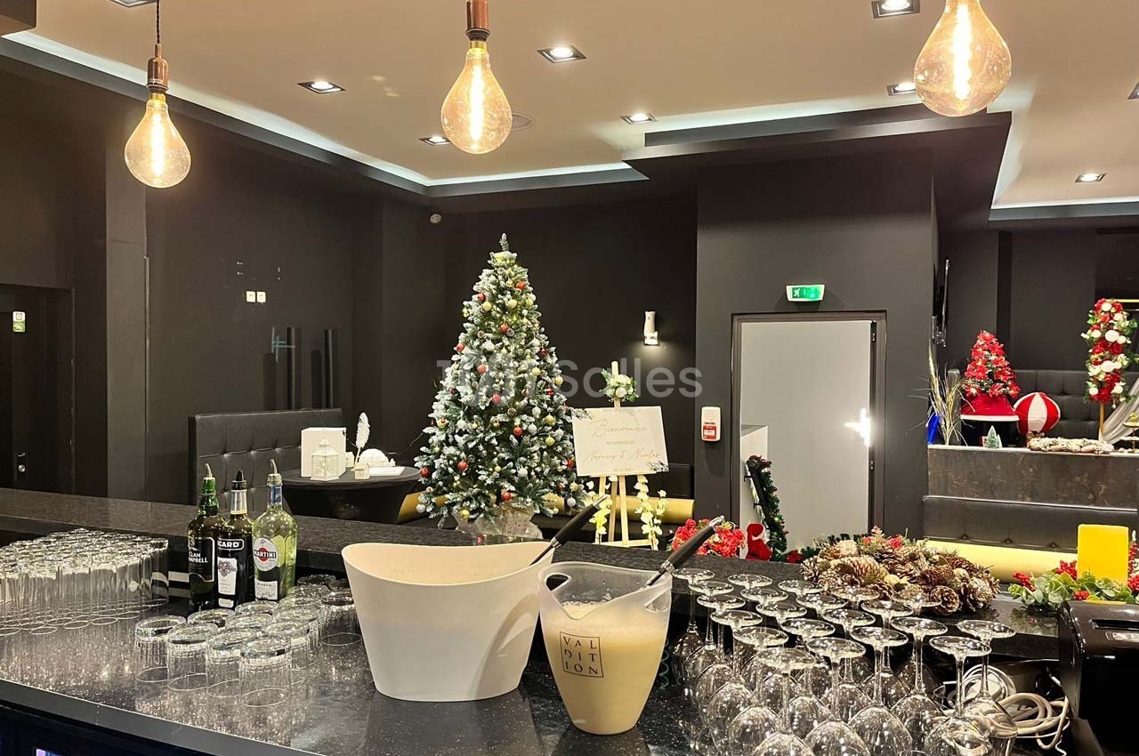Bar décoré pour Noël avec un sapin illuminé, des guirlandes, des verres alignés et des bouteilles sur le comptoir.