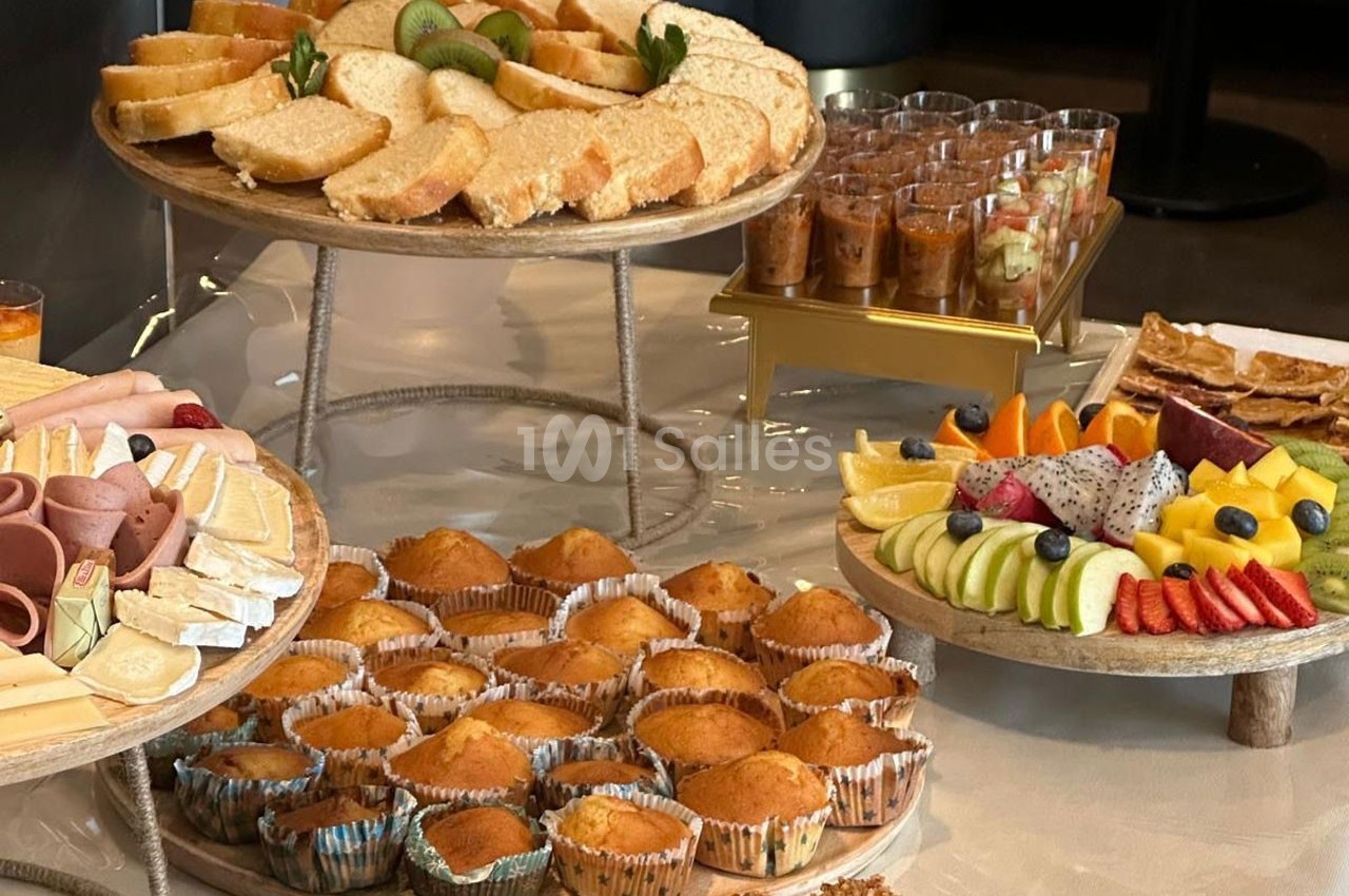 Plateaux garnis de muffins, fruits frais, pains, fromages et verrines, disposés sur une table pour un buffet.