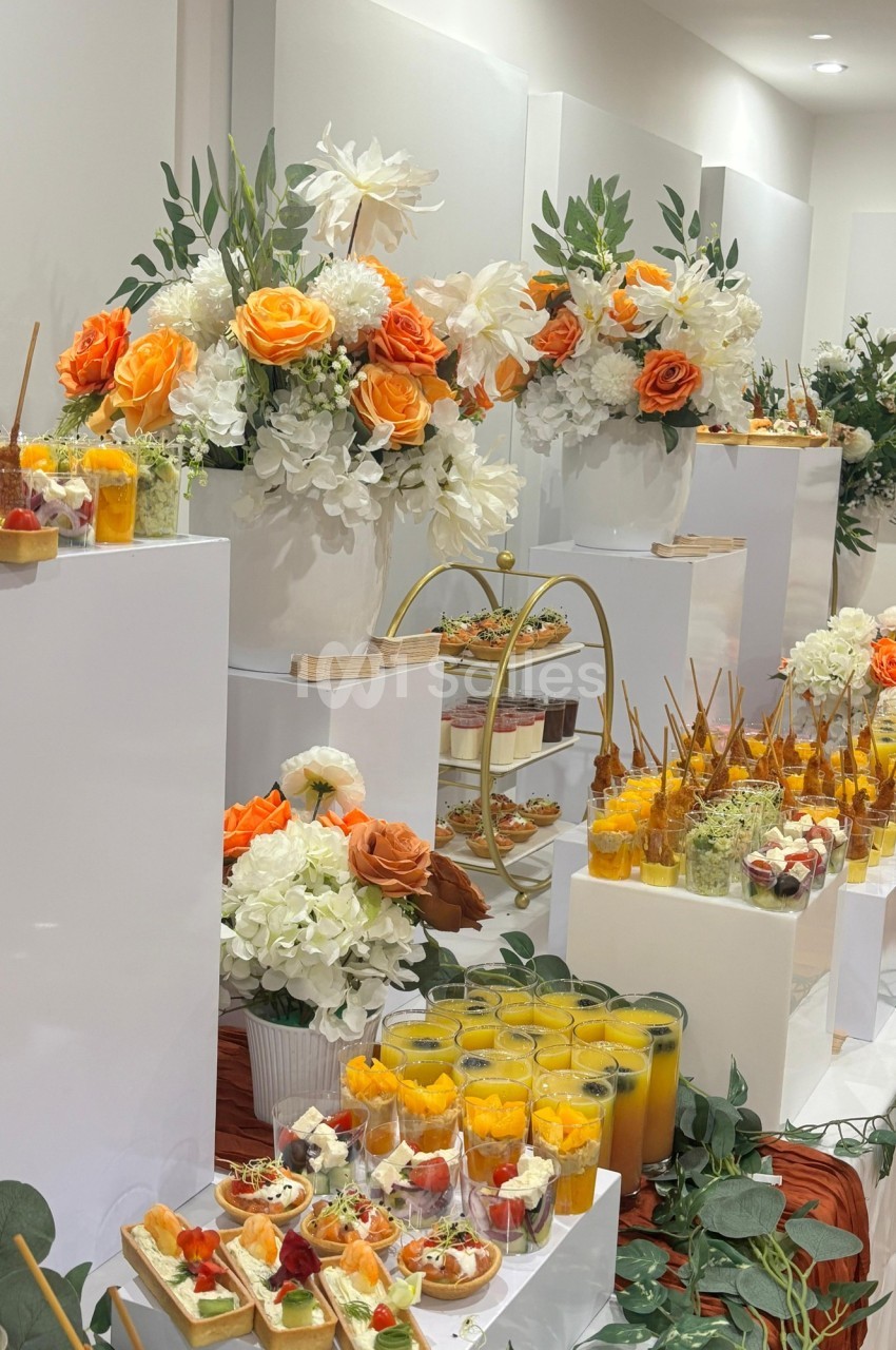 Buffet élégant avec fleurs orange et blanches, verrines, jus de fruits et amuse-bouches disposés sur des présentoirs blancs.