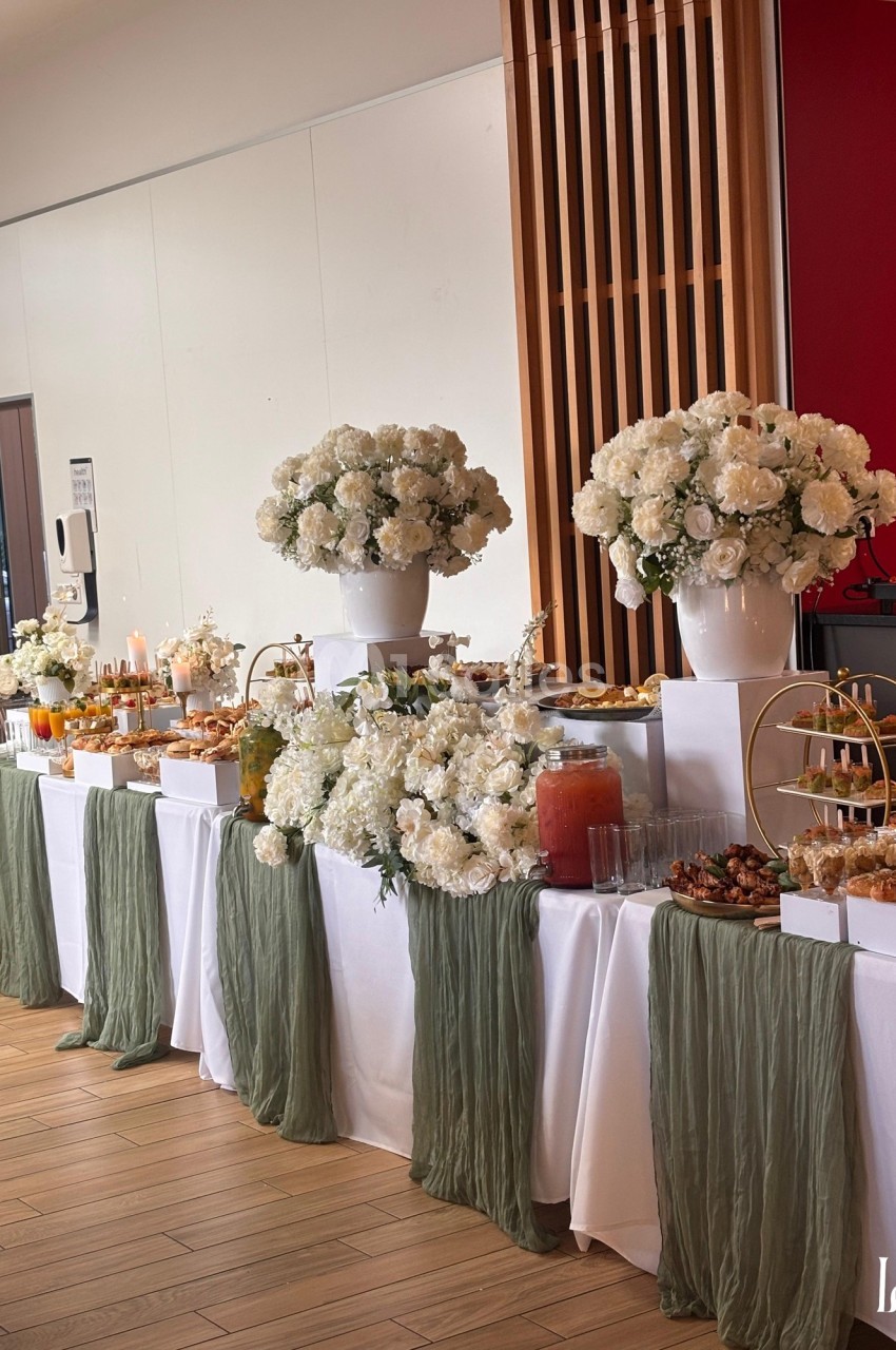 Table de buffet élégamment décorée avec des fleurs blanches, nappes vertes et divers plats et boissons disposés.