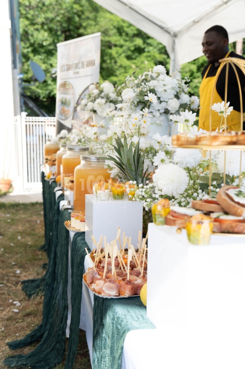 Buffet en extérieur avec boissons, fleurs blanches, fruits et amuse-bouches sur une table décorée de tissus verts.