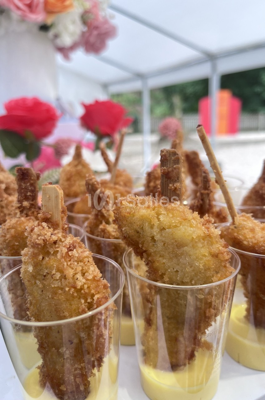Bouchées de poulet pané sur bâtonnets, servies dans des verrines avec une sauce jaune, décor floral en arrière-plan.