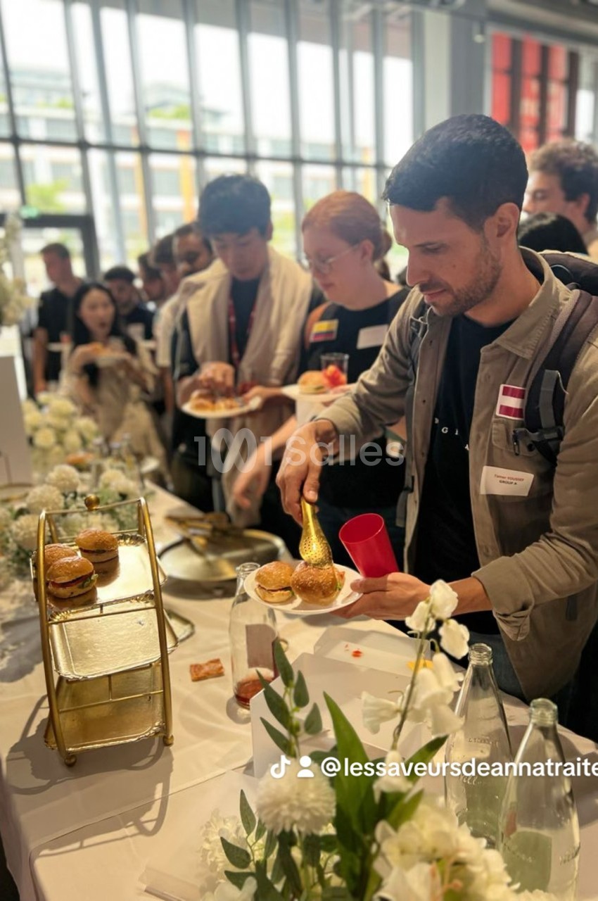 Des personnes se servent à un buffet décoré de fleurs blanches dans un espace lumineux avec de grandes fenêtres.
