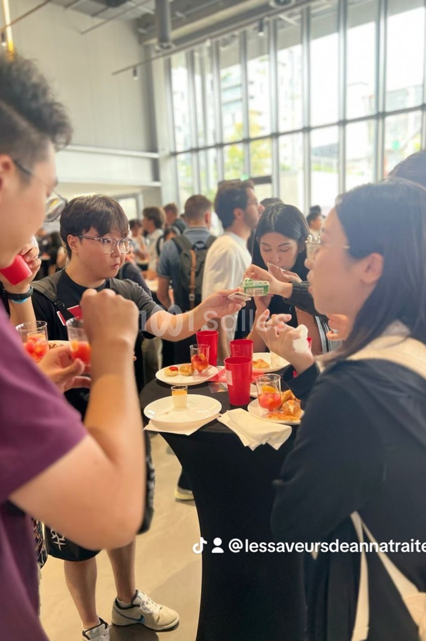 Des personnes debout autour de tables hautes dégustent des plats et boissons lors d'un événement convivial en intérieur.