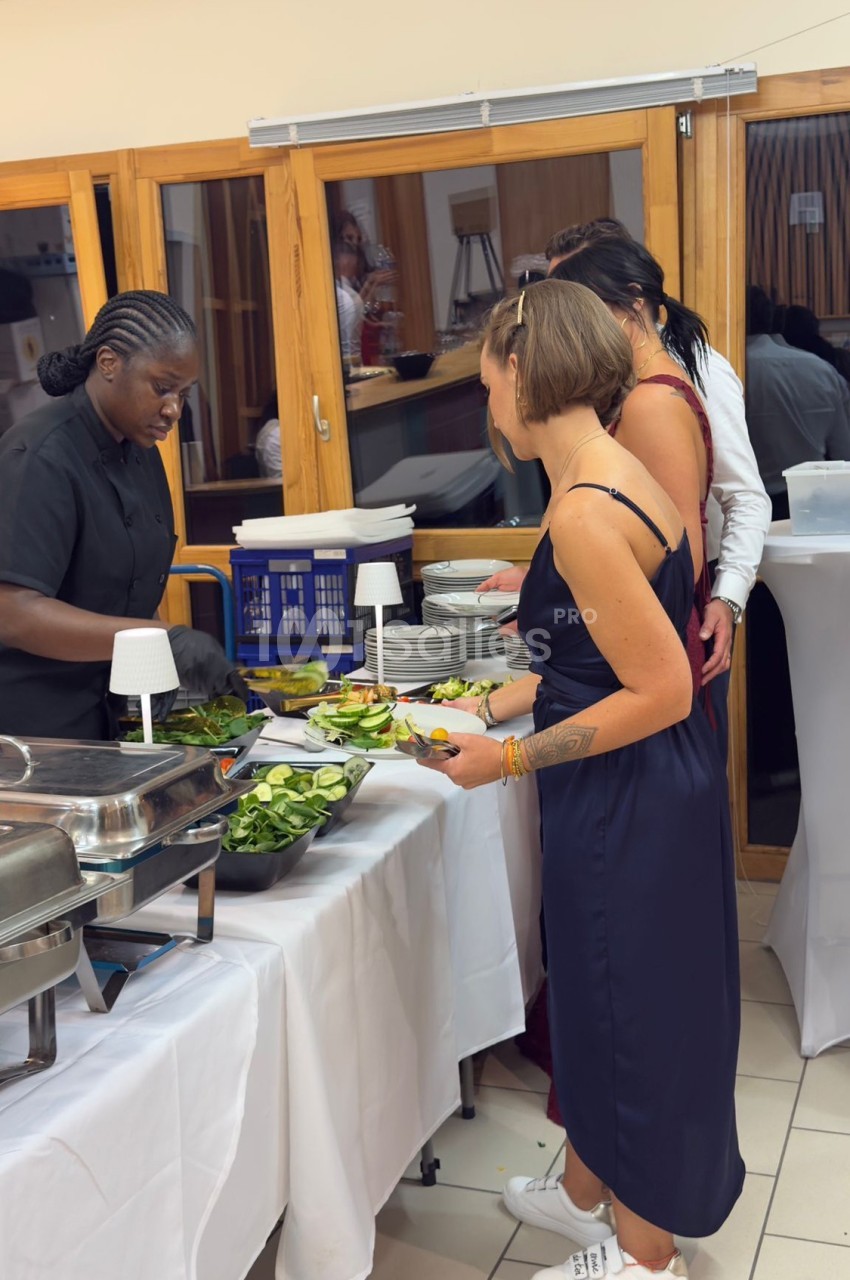 Une cuisinière sert des plats à des convives dans un buffet, avec des salades et des plats chauds disposés sur une table.