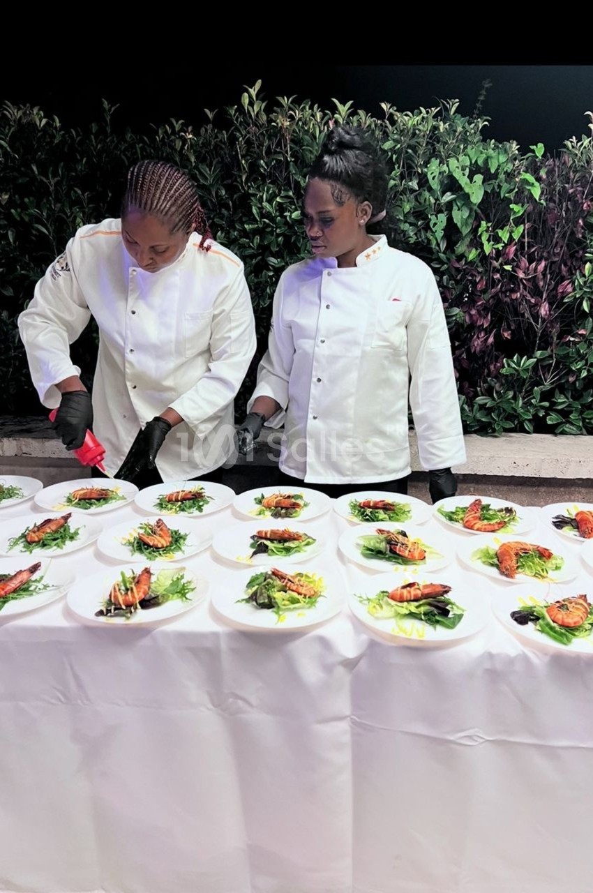 Deux chefs en tenue blanche dressent des assiettes garnies de légumes et de poisson sur une table blanche en extérieur.