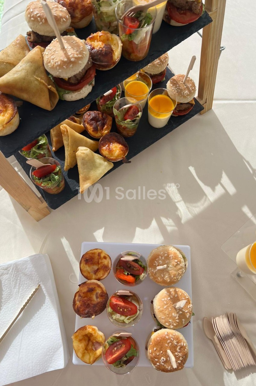 Plateau de mini-burgers, samoussas, quiches et verrines sucrées et salées disposés sur une table ensoleillée.