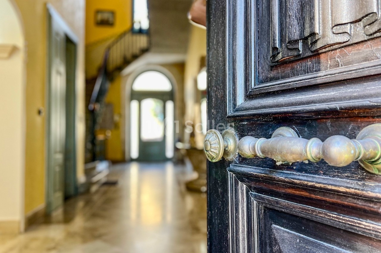 Porte en bois sombre entrouverte donnant sur un hall lumineux avec escalier et sol en marbre.