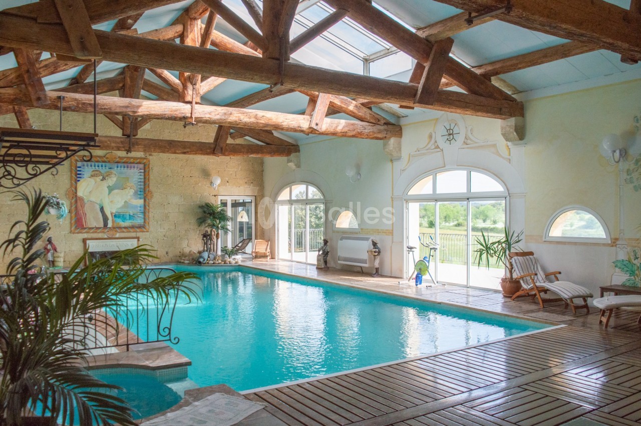 Piscine intérieure lumineuse avec charpente en bois, grandes baies vitrées et chaises longues en bordure.