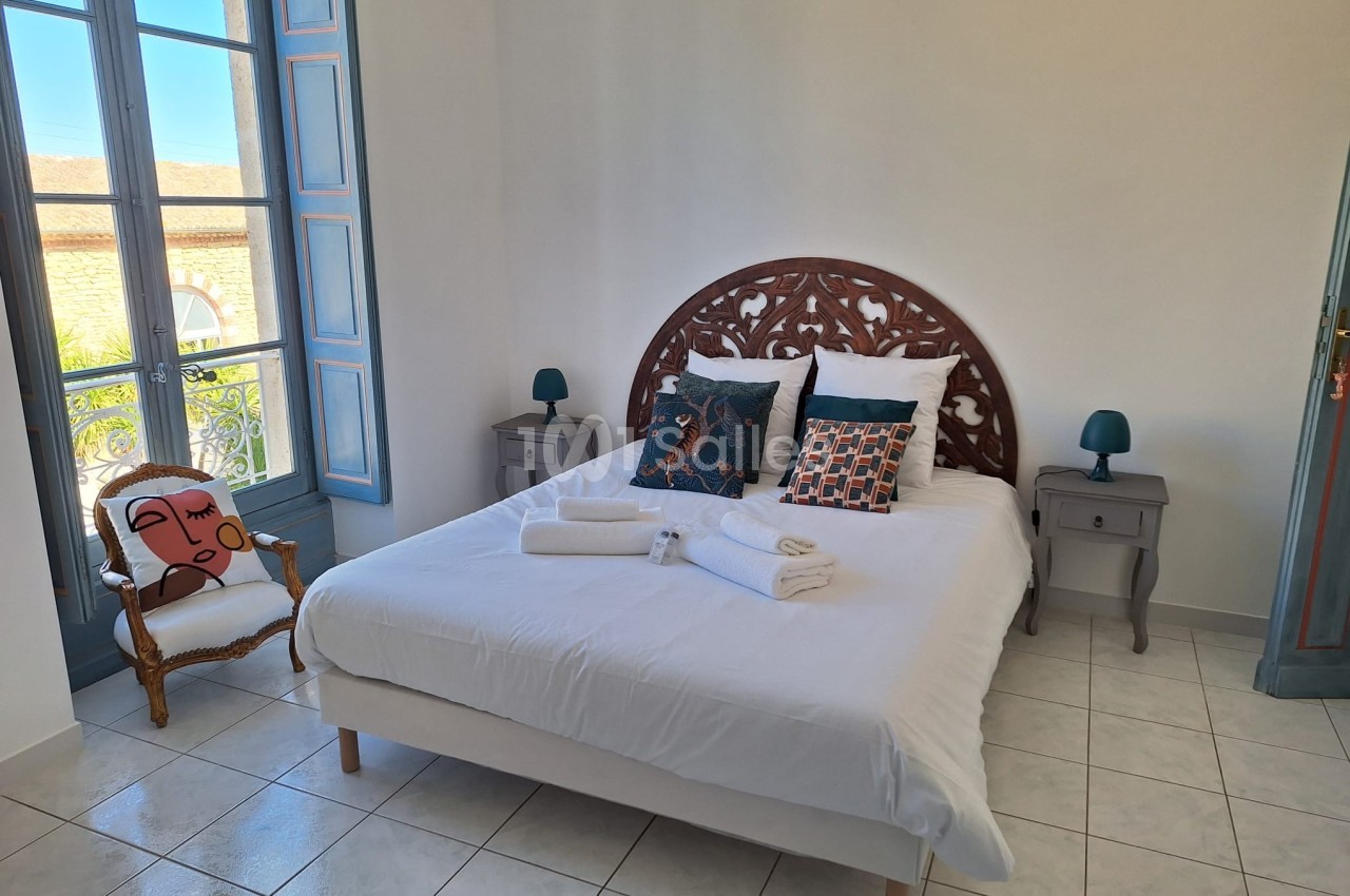 Chambre lumineuse avec lit double, tables de chevet, fauteuil et grande fenêtre donnant sur l'extérieur.