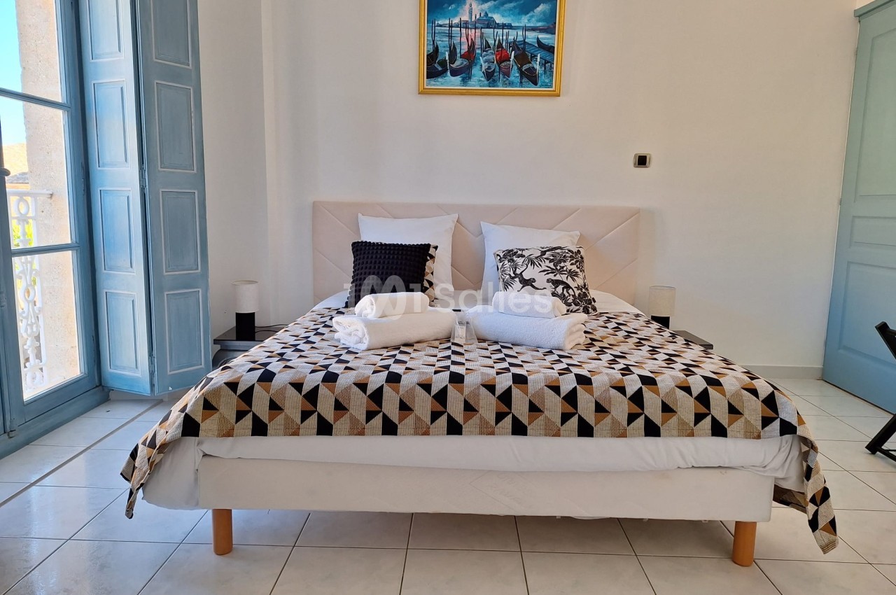 Lit double avec couverture géométrique, oreillers décoratifs et serviettes pliées, dans une chambre lumineuse avec balcon.
