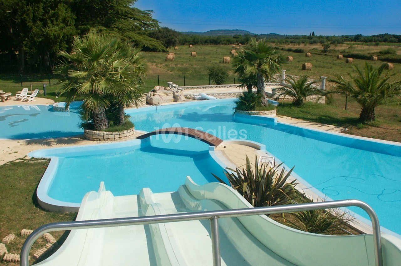 Piscine extérieure avec toboggans, pont en bois, palmiers et vue sur un paysage rural avec champs et bottes de foin.