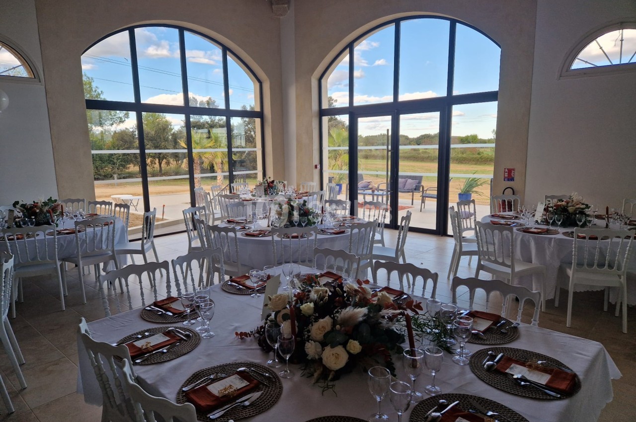 Salle de réception lumineuse avec tables décorées pour un événement, grandes baies vitrées donnant sur un paysage champêtre.