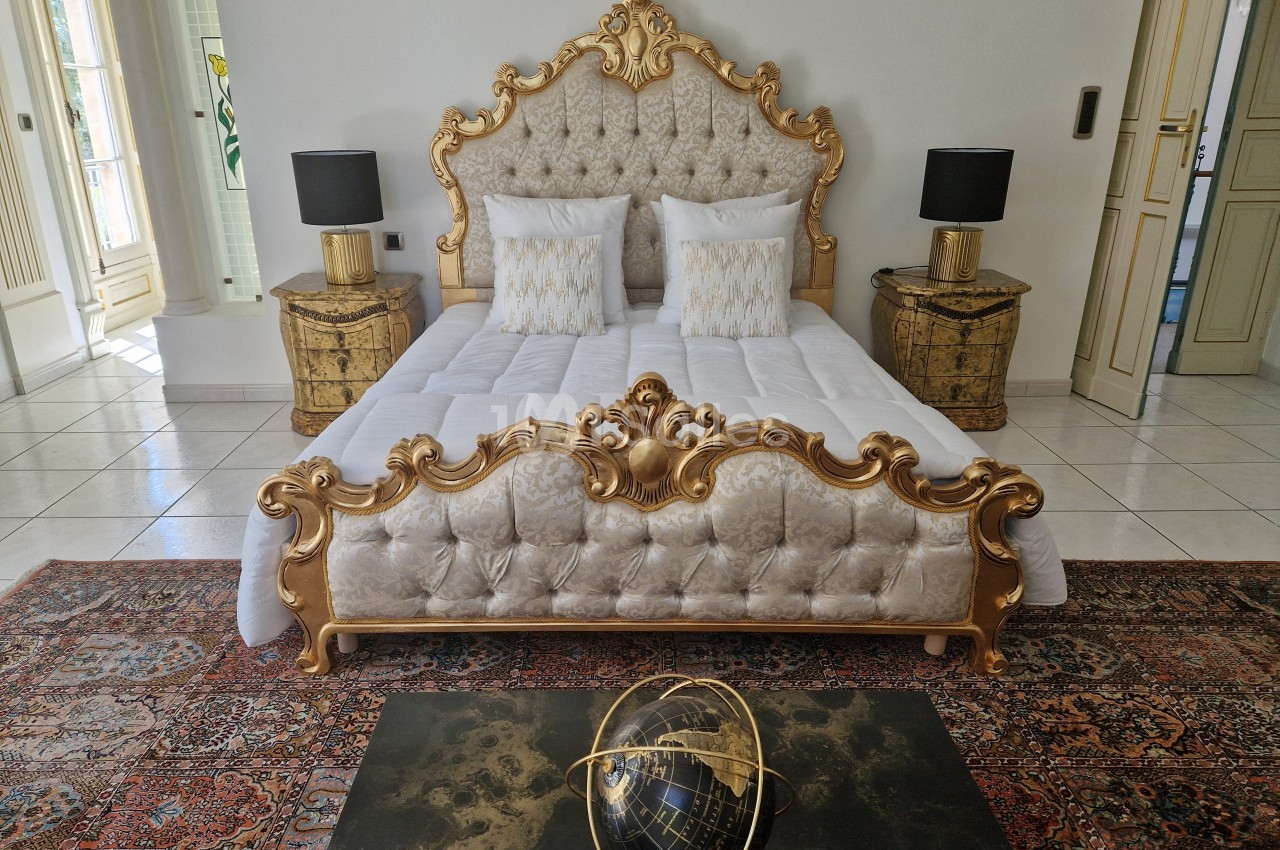 Lit à baldaquin doré avec tête de lit capitonnée, flanqué de tables de chevet et lampes, dans une chambre élégante.