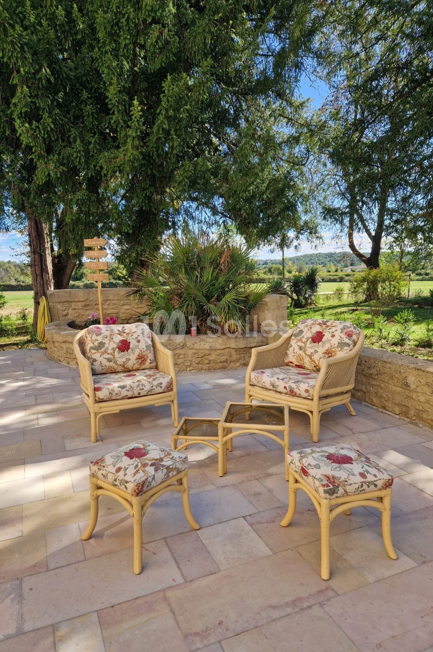 Mobilier en rotin avec coussins fleuris disposé sur une terrasse en pierre, entourée de verdure et d'arbres.