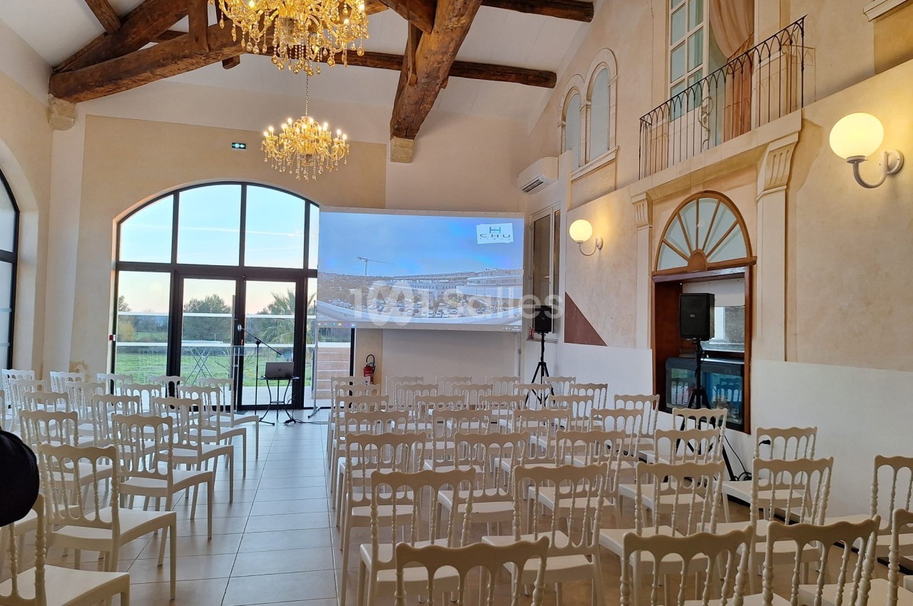 Salle lumineuse avec chaises blanches alignées, écran de projection allumé, poutres apparentes et lustres suspendus.