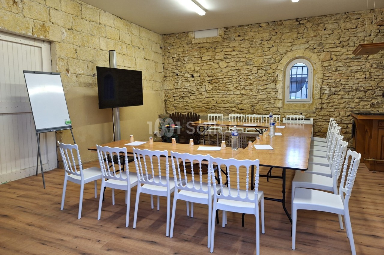 Salle de réunion avec murs en pierre, table rectangulaire entourée de chaises blanches, tableau blanc et écran.