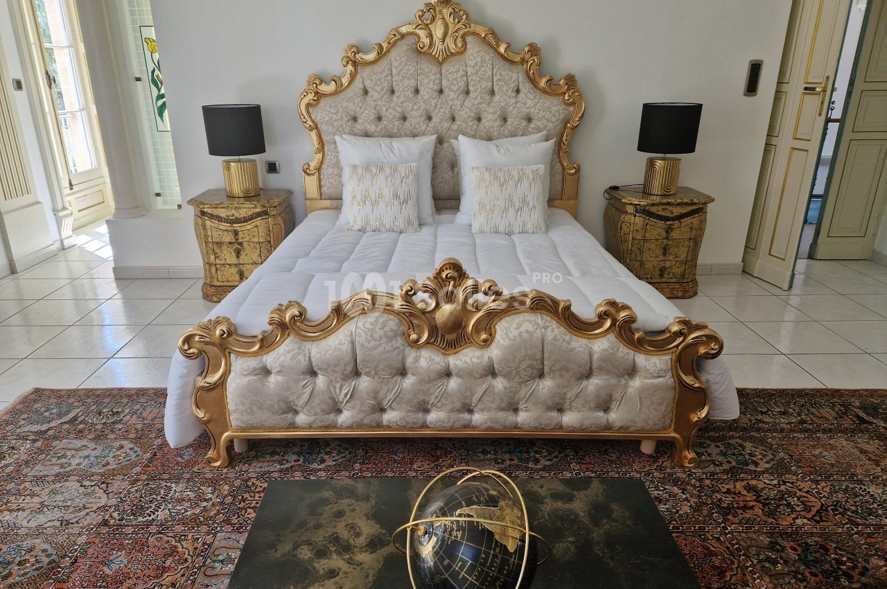 Lit à baldaquin doré avec tête de lit capitonnée, flanqué de tables de chevet et lampes, sur un tapis oriental.