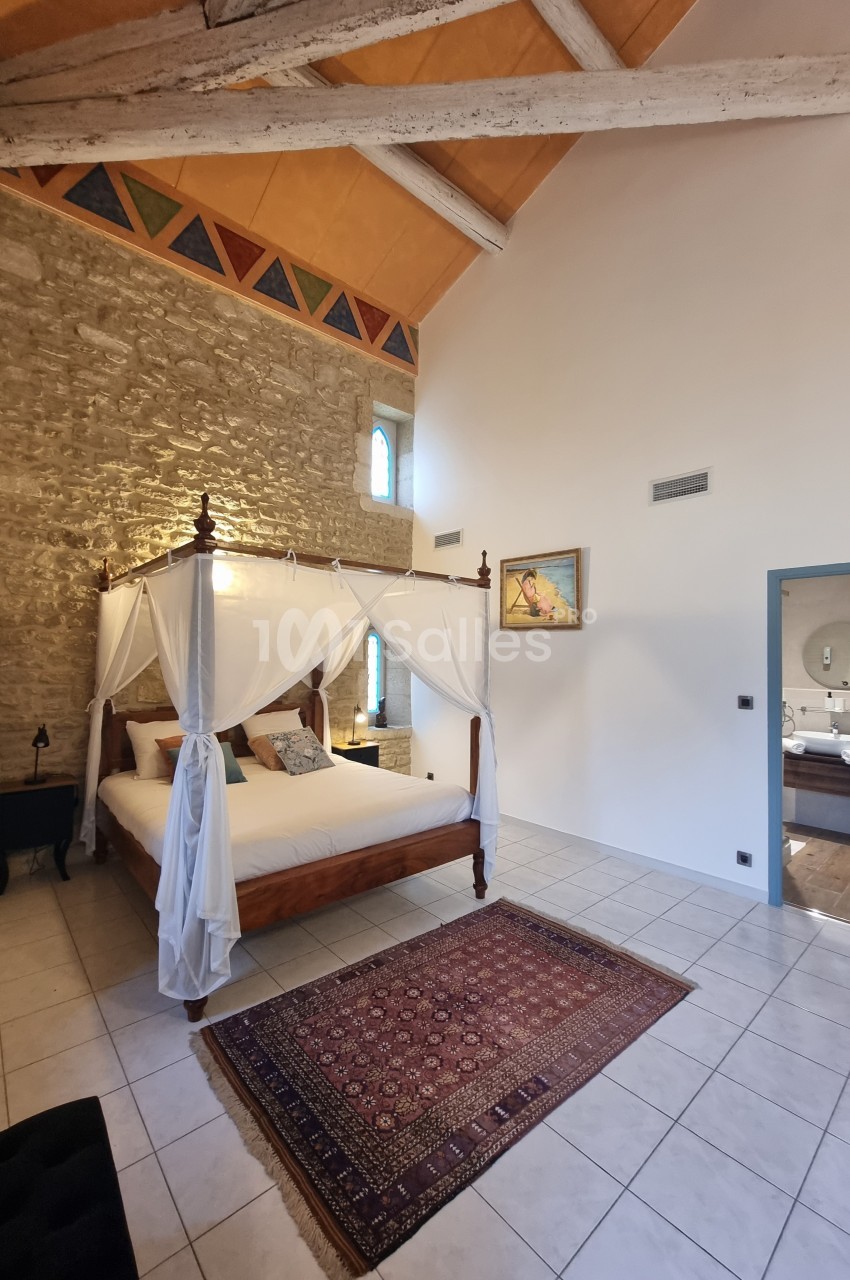 Chambre lumineuse avec lit à baldaquin, murs en pierre apparente, tapis coloré et plafond avec poutres en bois.