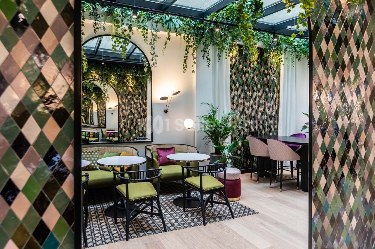 Espace intérieur avec tables, chaises, plantes suspendues et murs décorés de carreaux colorés en losanges.
