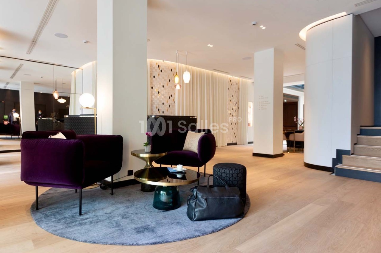 Espace salon moderne avec fauteuils violets, tables dorées, tapis rond gris et éclairage design dans un intérieur lumineux.