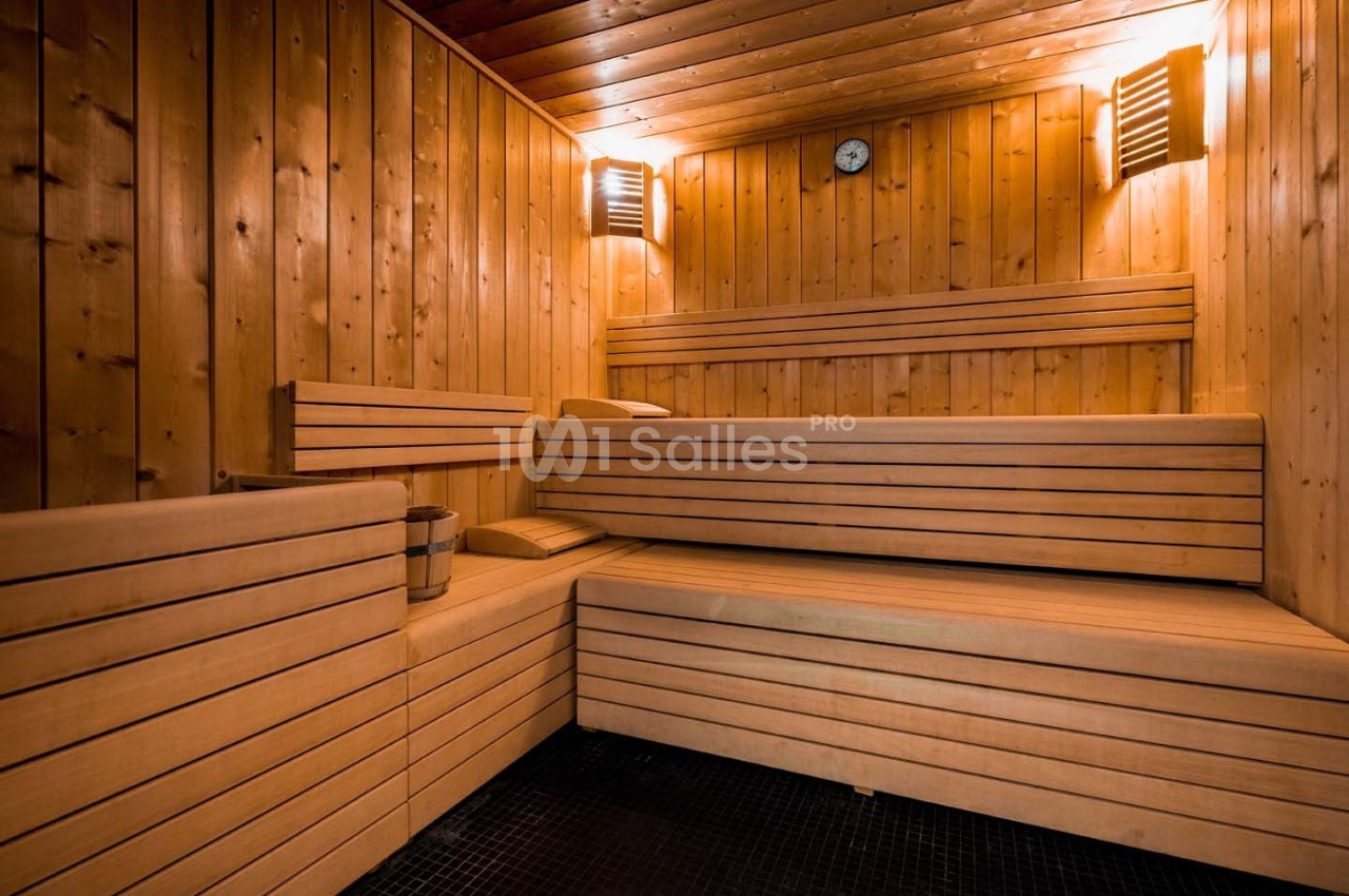 Intérieur d'un sauna en bois avec bancs en gradins, seau en métal et lumière tamisée.
